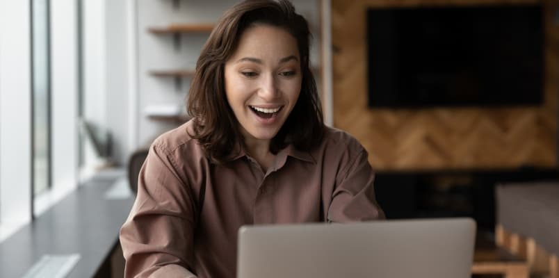 惊讶的女人笑着一个棕色的衬衫,她坐在桌子上,用她的笔记本电脑。
