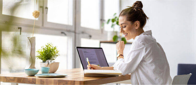 一个微笑的女人坐在她的办公桌前，拿着笔记本电脑，写着一些笔记。